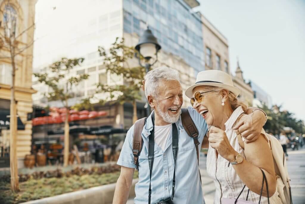Seguro viagem anual vale a pena? Para quem faz mais de uma viagem por ano, a conta fecha 3 Casal de idosos turistando em uma cidade. Eles estão sorridentes.