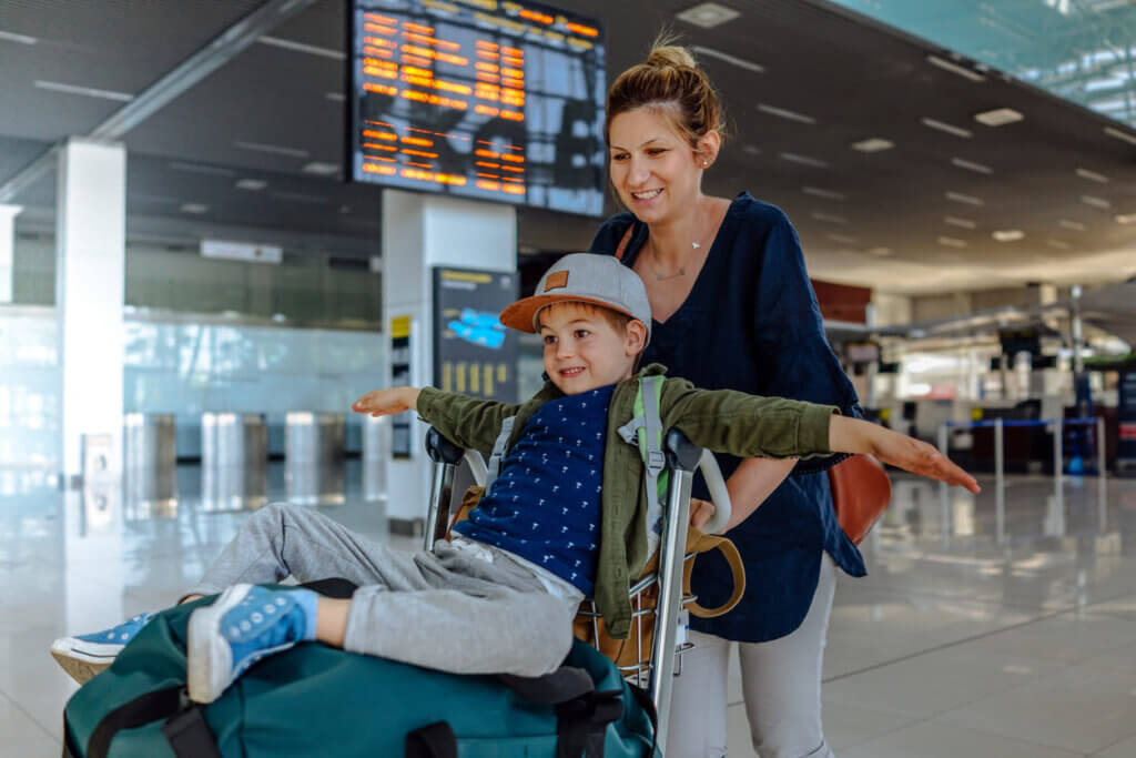 Seguro viagem anual vale a pena? Para quem faz mais de uma viagem por ano, a conta fecha 2 uma mulher com uma criança no aeroporto. Há malas no carrinho e o menino está em cima delas.