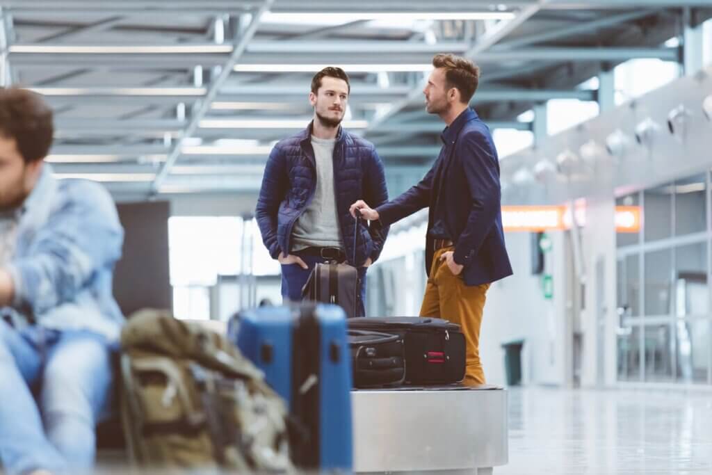 Mala extraviada: o que fazer para localizar a bagagem e acionar o seguro 2 Dois homens conversando próximos à esteira de malas do aeroporto enquanto aguardam suas bagagens.