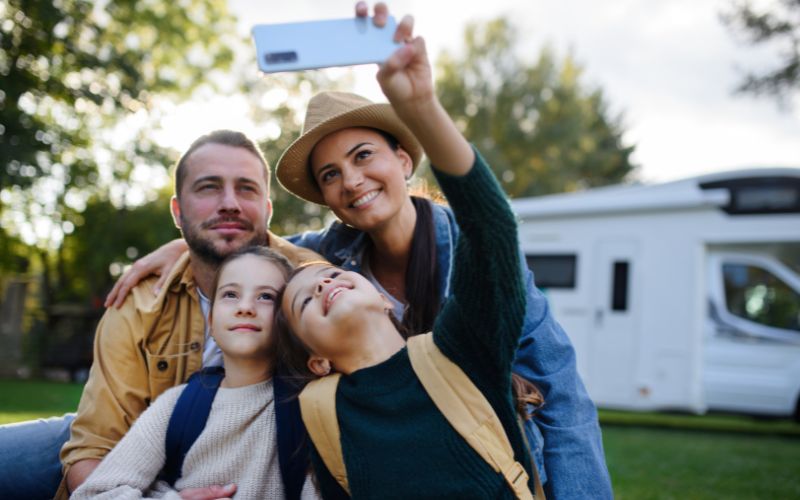 Viagem de Família com as Crianças para Destinos Divertidos 3 Família jovem feliz com dois filhos tirando selfie com caravana ao fundo ao ar livre.