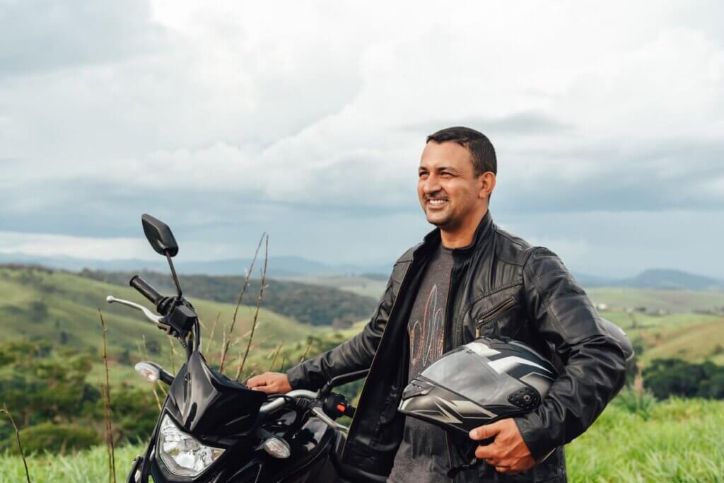 Seguro viagem de moto: o que é, o que cobre e por que é diferente do seguro comum 3 Um homem sorridente parado na estrada, em pé e encostado na moto, com o capacete na mão. Ao fundo há uma paisagem de montanhas com céu carregado de nuvens.