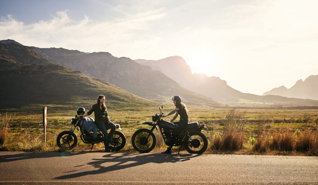Seguro viagem de moto: o que é, o que cobre e por que é diferente do seguro comum 1 Casal de motoqueiros parados na estrada, no acostamento, em dia ilumidado pelo sol com montanhas ao fundo.