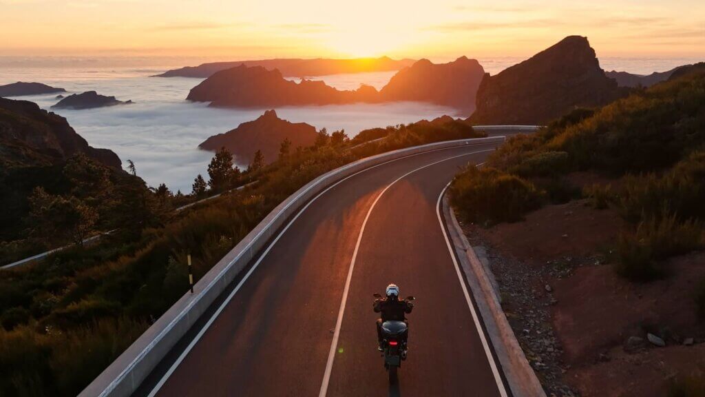 Seguro viagem de moto: o que é, o que cobre e por que é diferente do seguro comum 2 Um pessoa dirigindo a moto na estrada com vista deslumbrante do pôr do sol com montanhas e nuvens ao redor.