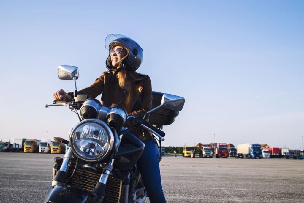 Seguro viagem de moto: o que é, o que cobre e por que é diferente do seguro comum 4 Uma mulher sentada em uma moto e sorridente. Céu limpo ao fundo e vários caminhões estacionados.