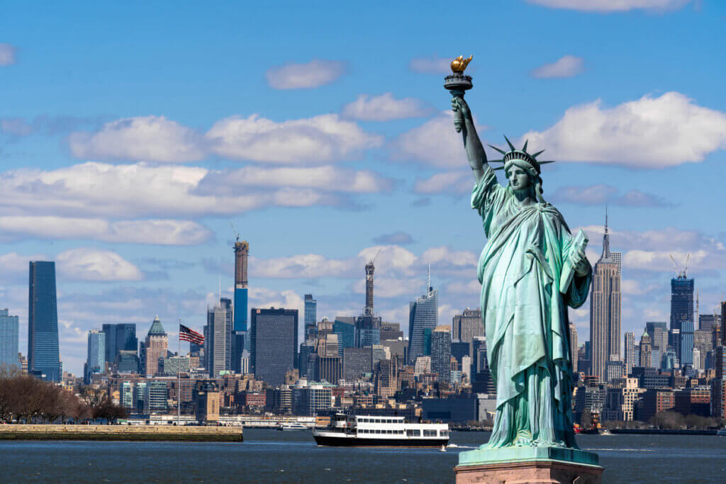 Imagem da Estátua da Liberdade em Nova York com a linha do horizonte da cidade ao fundo sob céu parcialmente nublado.