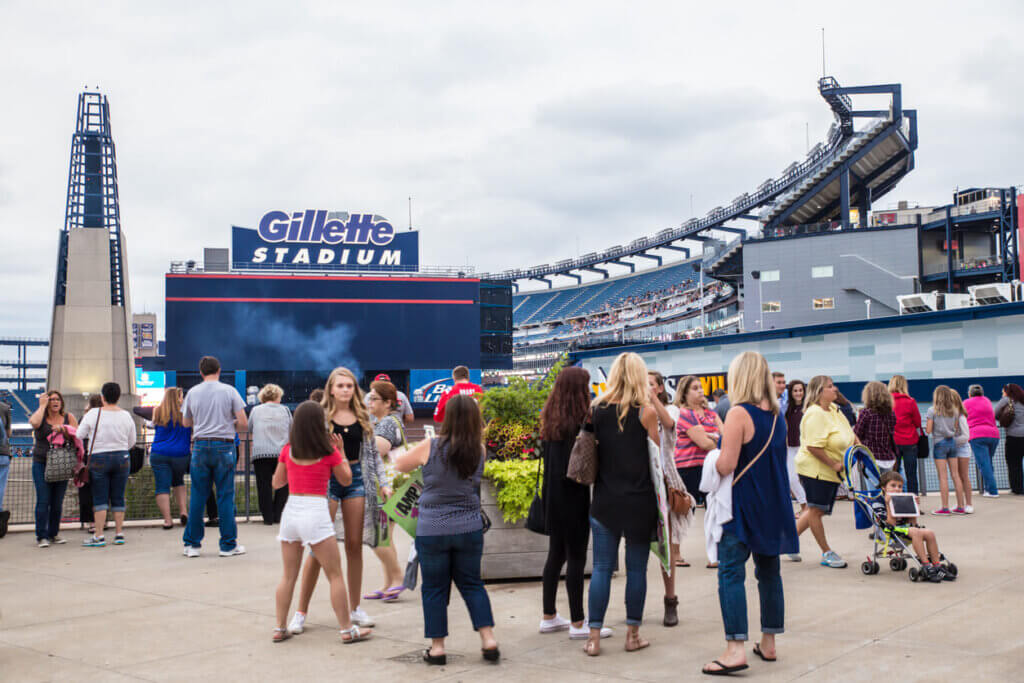 Viaje para Boston para a Copa do Mundo de Futebol 2026 2 Visitantes no estádio Gillette Stadium, em Boston, participando de um evento ao ar livre com pessoas de diferentes idades, ao redor de um grande telão e estruturas do estádio ao fundo.