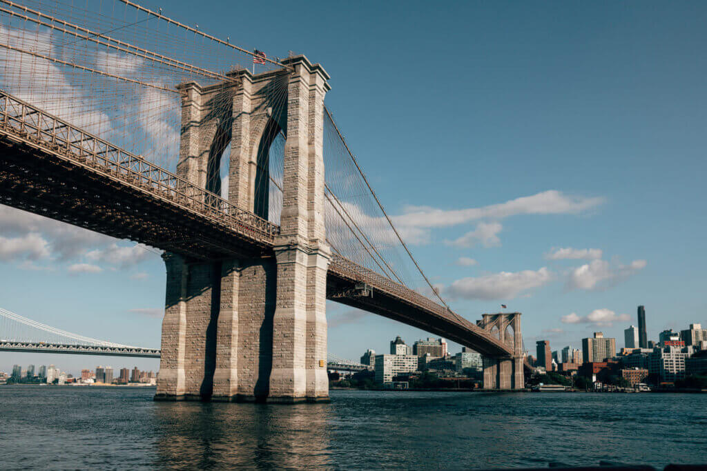Imagem do Brooklyn Bridge em Nova York, destacando sua estrutura de stonework e vista para o rio com prédios ao fundo em um dia claro.