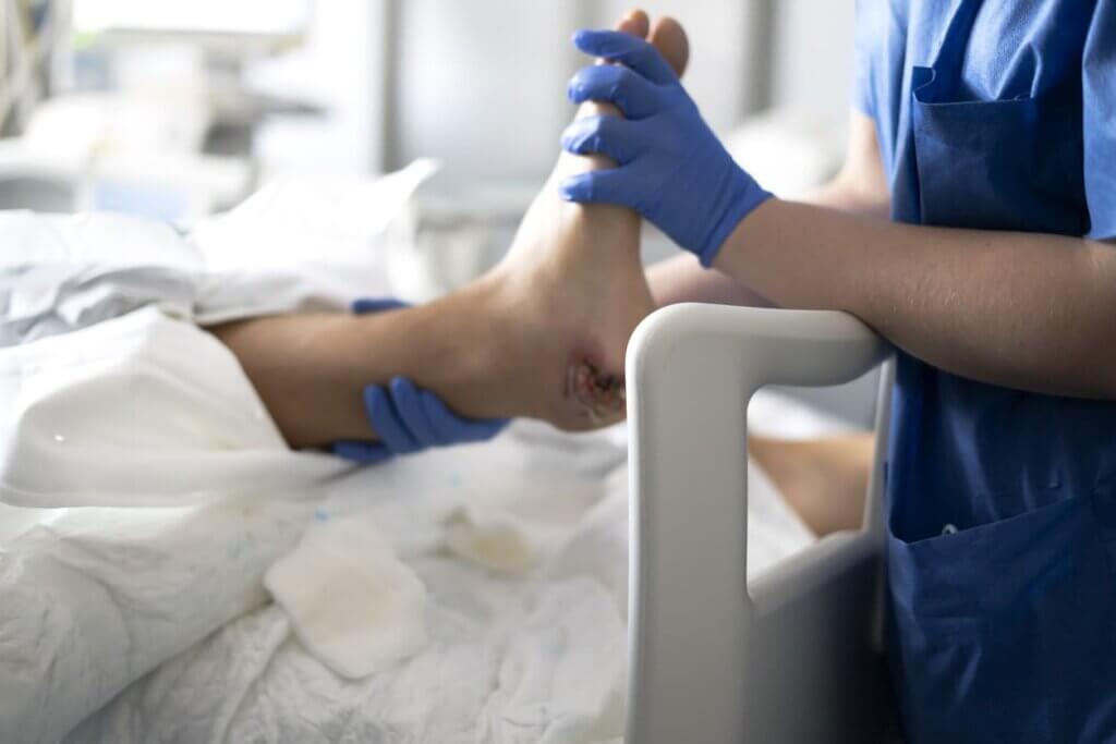 Profissional de saúde realiza exame ou procedimento no pé de um paciente em ambiente hospitalar, com uso de luvas azuis e vestuário hospitalar.