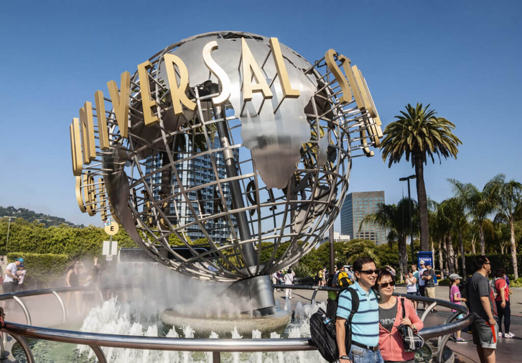 Fachada da fonte do globo da Universal Studios em Los Angeles com visitantes sorridentes na frente, árvores e céu azul ao fundo.