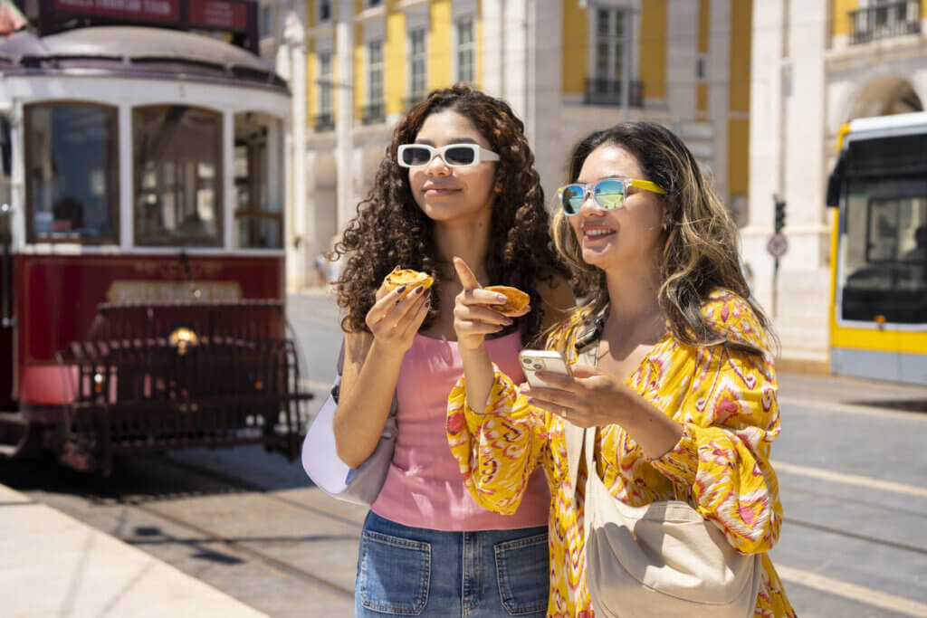 Quanto custa uma viagem para Portugal: orçamento detalhado por categoria 4 Turistas comendo pastel de nata em Lisboa com bondinho desfocado ao fundo