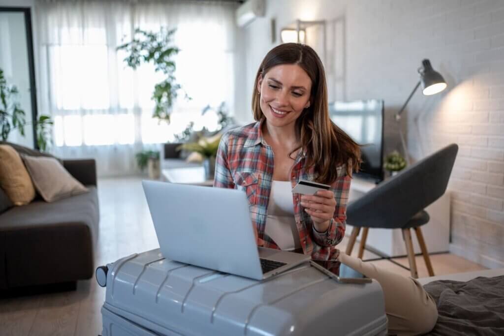 Viajar de avião pela primeira vez: guia completo para iniciantes em 2026 2 Mulher jovem na sala de casa mexendo em um notebook cinza que está apoiado na mala de viagem da cor cinza. Ela segura um cartão de crédito na mão com a intenção de realizar uma compra.