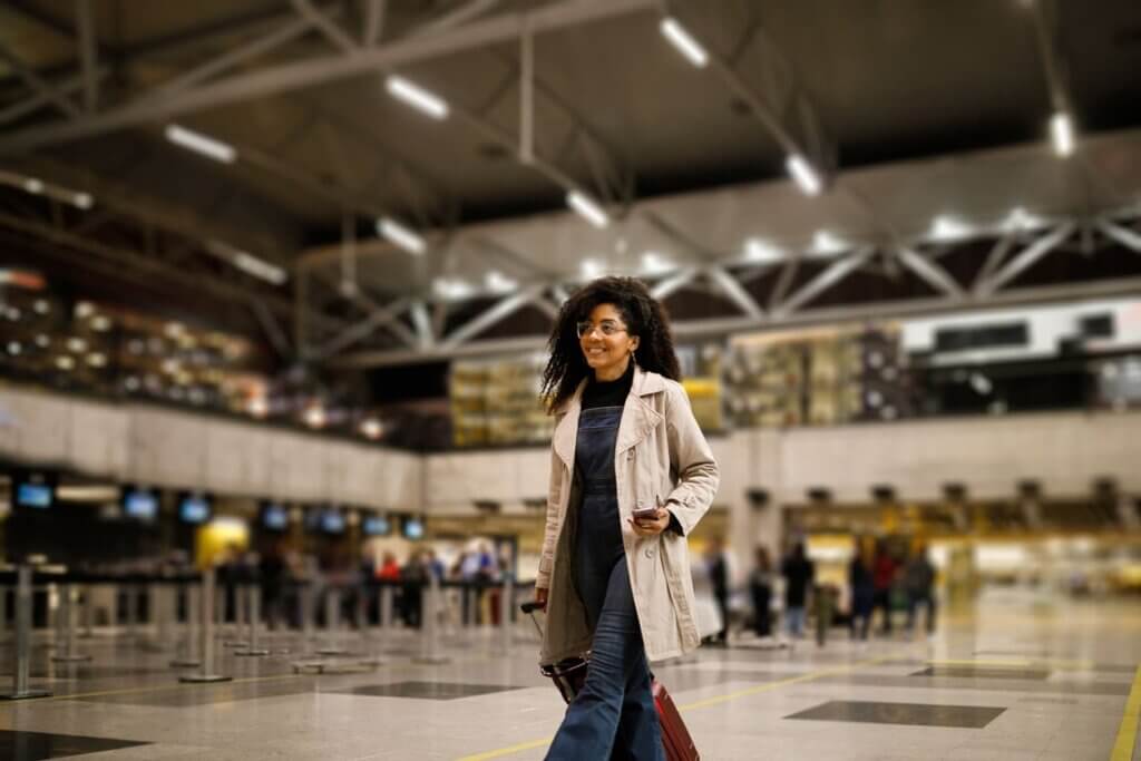 Viajar de avião pela primeira vez: guia completo para iniciantes em 2026 4 Mulher jovem andando no aeroporto com a sua bagagem de mão.