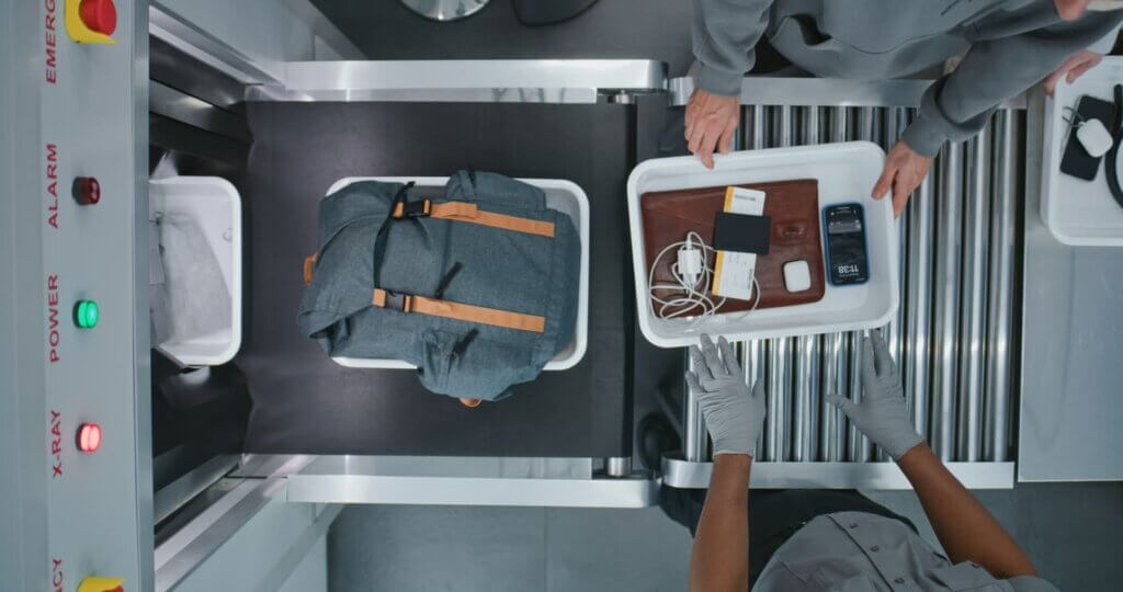 Checklist para viajar completo para viagens nacionais e internacionais 4 Vista superior de diversas pessoas, passageiros colocando itens pessoais em bandejas plásticas para procedimentos de triagem