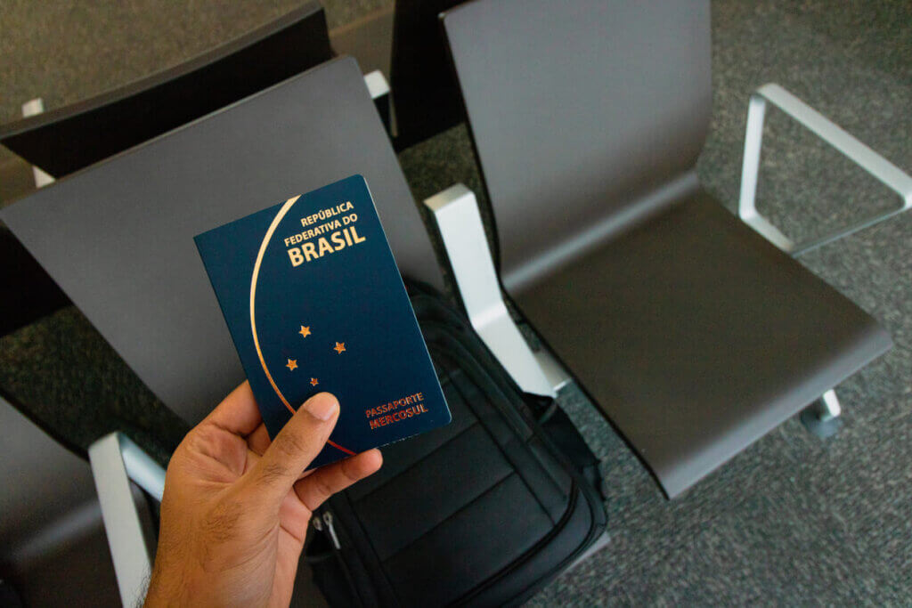 Viajar de avião pela primeira vez: guia completo para iniciantes em 2026 3 Mão de um homem segurando o passaporte brasileiro. Ao fundo da foto está uma mochila preta em uma cadeira da sala de espera do aeroporto.