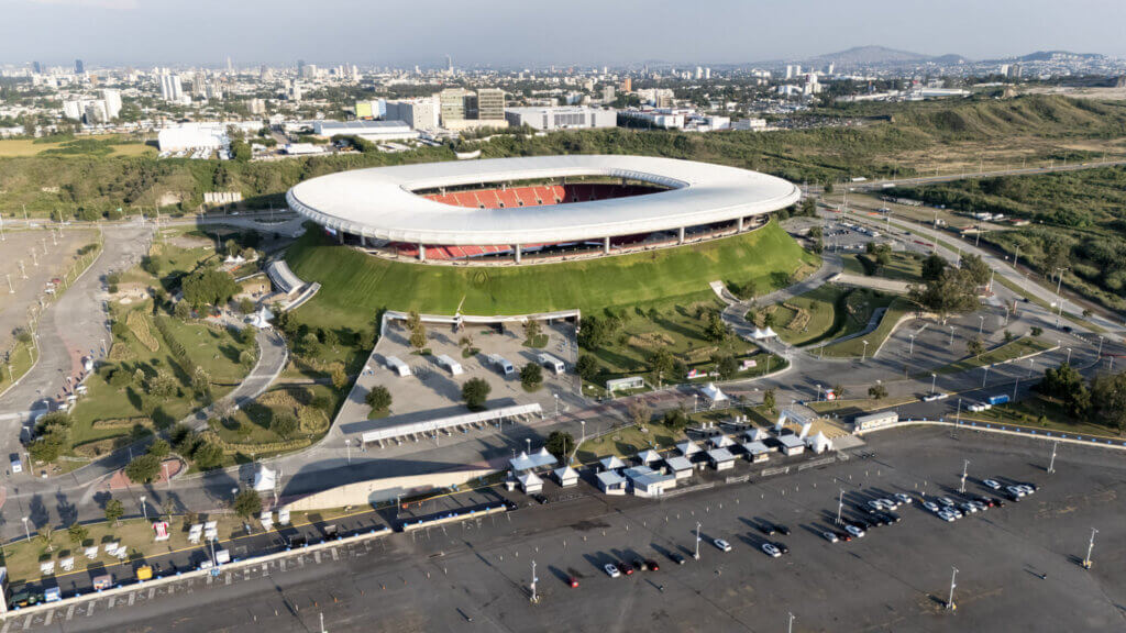 Vista aérea do estádio guadalajara em dia ensolarado com estacionamento e carros na frente.