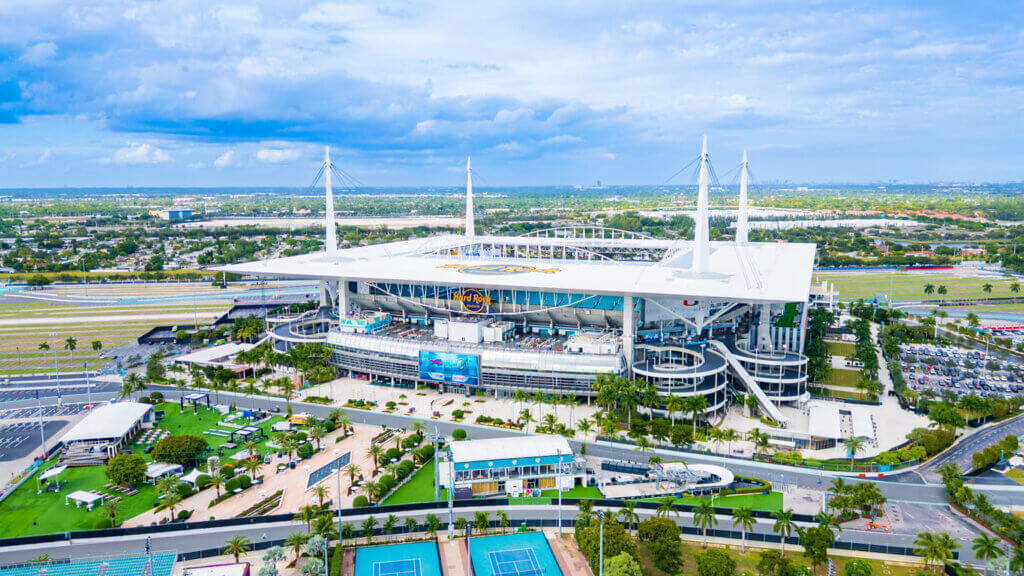Imagem aérea do Hard Rock Stadium em Miami, um estádio moderno e gigante, com estrutura arquitetônica impressionante, cercado por áreas verdes e pistas de corrida.