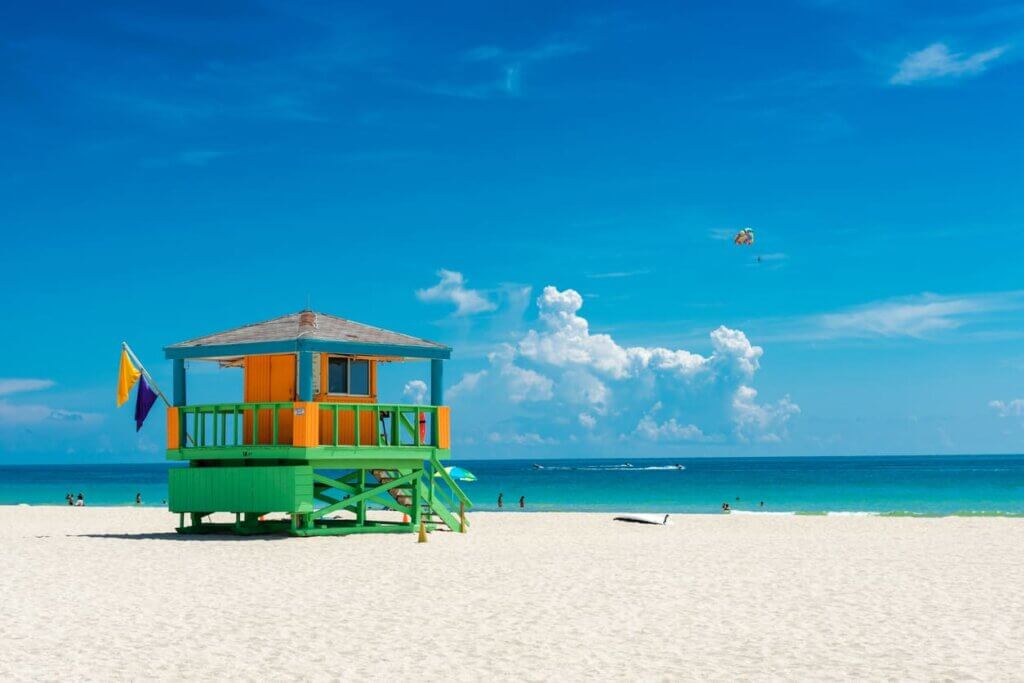 Casal de salva-vidas colorido na praia de areia branca com céu azul, nuvens e mar cristalino ao fundo em South Beach, Miami. 