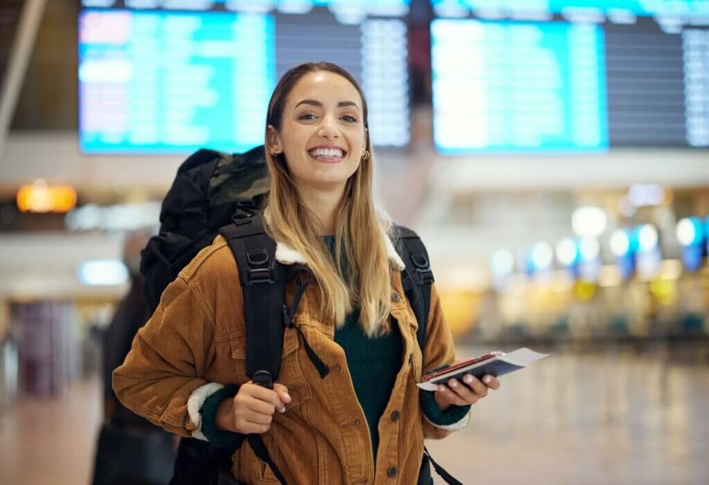 Custo de viagem internacional em 2026: como planejar cada despesa do seu roteiro 2 Jovem mulher sorridente segurando um celular e com painel de voos ao fundo do aeroporto