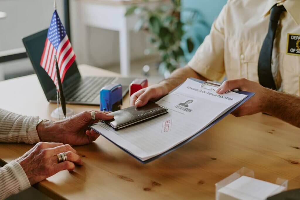 Pessoa entregando documentos e carteira para o oficial de segurança no balcão de um escritório, com bandeira dos EUA e laptop ao fundo, simbolizando controle de identidade.