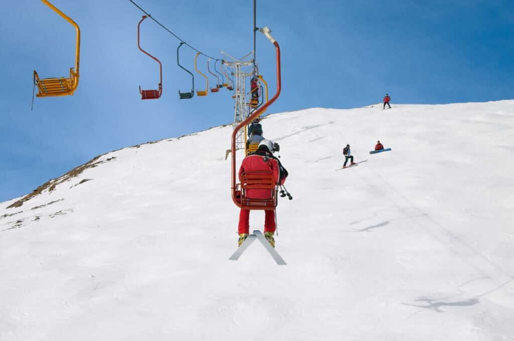 Pessoas esquiando na neve em uma estação de montanha, com cadeira do teleférico ao centro e vários esquiadores ao fundo em uma pista branca sob céu azul