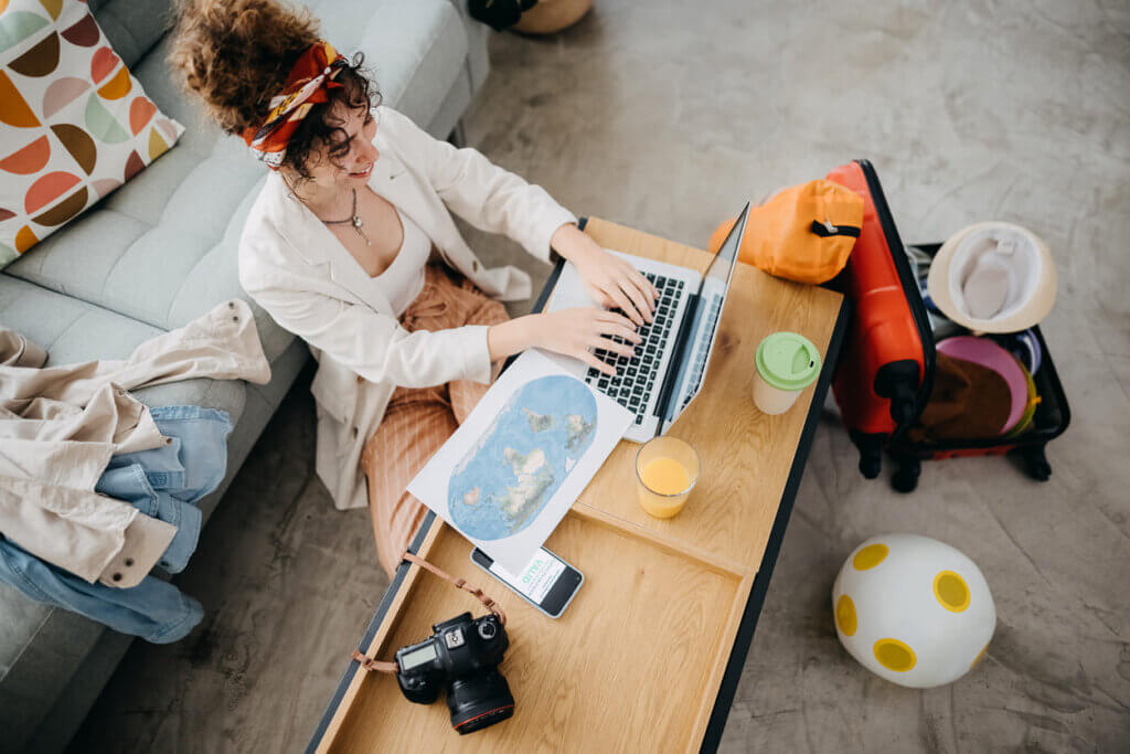 Como funciona o seguro viagem: da contratação ao acionamento, tudo o que você precisa saber 4 Foto tirada de cima de uma mulher sentada no chão de sua casa e encostada no sofá enquanto usa um notebook para planejar a sua viagem