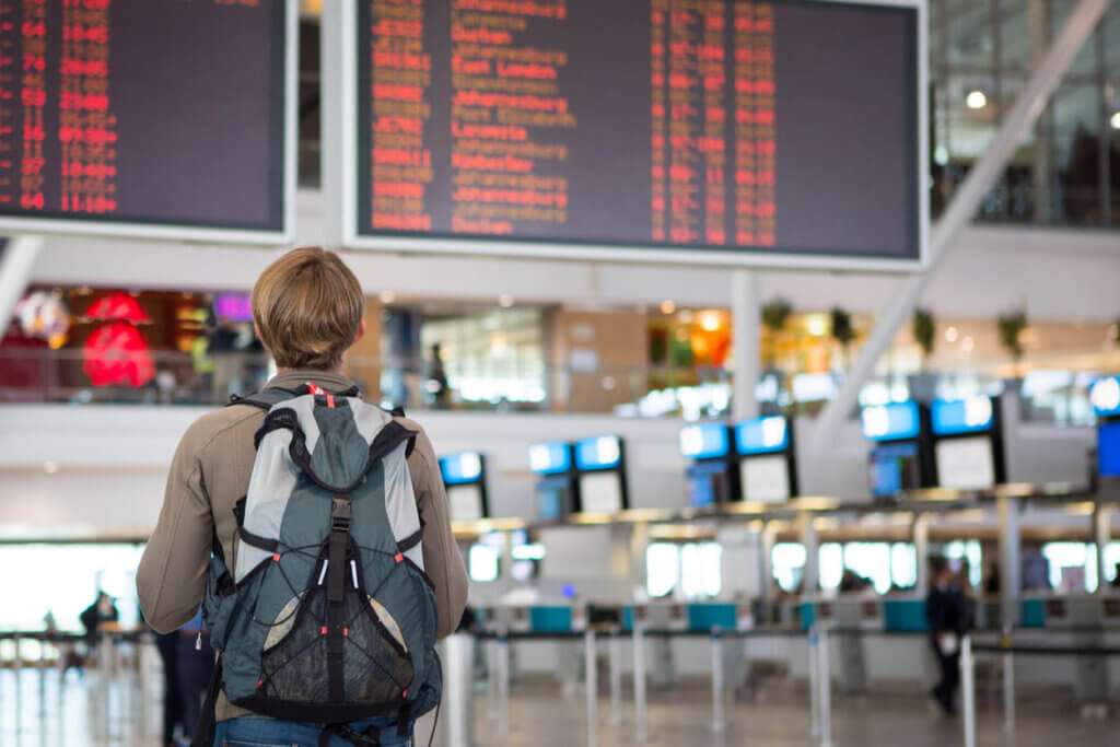 Como funciona o seguro viagem: da contratação ao acionamento, tudo o que você precisa saber 2 Um pessoa com mochila nas costas olhando para telas sobre status de voos no aeroporto