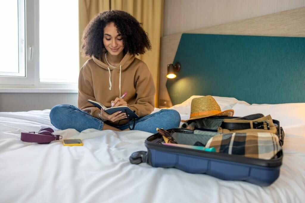 Diferença entre seguro saúde e seguro viagem: o que é cada um e qual contratar para viajar 2 Mulher sentada na cama com mala de viagem aberta e fazendo anotações em um caderno.