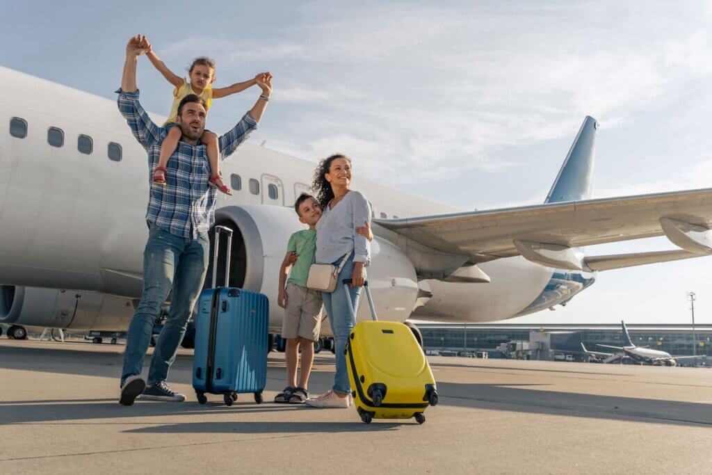 Diferença entre seguro saúde e seguro viagem: o que é cada um e qual contratar para viajar 1 Pais felizes e seus filhos esperando para viajar juntos com um avião estacionado ao fundo