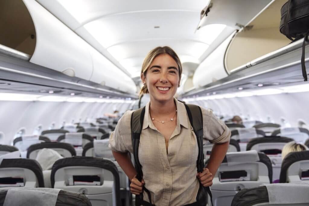 Mulher jovem com mochila nas costas sorrindo para foto no corredor de uma aeronave