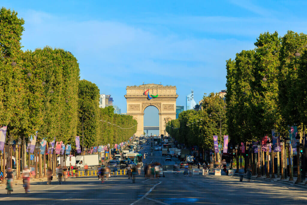Imagem da avenida com o Arco do Triunfo, em Paris, ao fundo. Árvores ao lado e dia com céu azul.
